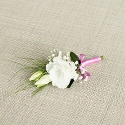 A finely detailed wedding boutonnière rests diagonally across a softly lit, oatmeal-coloured fabric that resembles natural linen, its woven threads giving a warm, homely texture reminiscent of table linens seen in Queensbury's cosy cafes. The focus of the arrangement is a single, impeccable white lisianthus flower, fully open and slightly off-centre to the right. Its ruffled petals spiral in gentle layers, silky and luminous, creating a sense of quiet elegance and calm. Nestled beside it, on the lower left, are two elongated eustoma buds in a tender mix of ivory and pale green, still closed and smooth, hinting at the unfolding of a special day, like a couple just about to say their vows at a Queensbury church or registry office. Scattered among these main blooms are dainty clusters of gypsophila, or baby's breath: countless tiny white flowers that create a cloud-like halo, softening the edges and adding a touch of airy romance. From behind the flowers, slender, dark green, grass-like leaves fan out gracefully toward the left, lightly arching as if caught by a breeze across Queensbury Park. A single broader, deeper green leaf sits under the white bloom, giving the design a sense of balance and structure. At the lower right, the stems are cleanly trimmed and wrapped with a vivid fuchsia satin ribbon, tied into a small, neatly formed bow whose bright pink sheen contrasts beautifully with the neutral background and crisp white petals. The gentle, even lighting allows every texture-from smooth ribbon to velvety petal-to be clearly seen, evoking the feeling of a carefully prepared buttonhole waiting on a dresser before being pinned to the lapel of a groom, best man, or prom guest ready to head out into the streets of Queensbury for a day of celebration and photographs.