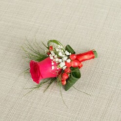 A finely crafted boutonniere is displayed on a pale beige, lightly textured fabric that resembles natural linen, the kind you might find dressing reception tables at a cosy Queensbury hotel or local function room. The composition is anchored by a single hot fuchsia rose, its petals tightly layered at the centre and gently opening outwards, each velvety curve full of colour and quiet drama. Clustered at the base of the rose are delicate sprays of white baby's breath, their tiny star-like blooms forming a foamy ring that softens the strong pink and adds a sense of innocence and tradition. Tucked among these white blossoms sit rounded red hypericum berries, polished and luminous, like bright beads plucked from hedges in nearby parks, bringing depth and a friendly hint of festivity. Around the floral heart, rich green foliage has been expertly arranged: broader, darker leaves are curled and shaped into sleek arcs that frame the rose, while finer, feathery fronds extend outwards to give the buttonhole lightness and movement. The stems are tightly wrapped in a luxurious red satin ribbon, the fabric smooth and slightly lustrous, finished in a neat, compact knot that ensures comfort when pinned to a lapel. Gentle, diffuse light washes over the scene, emphasising the contrast between soft petals, glossy berries, and the woven background, so that every detail of the florist's handiwork is visible. This elegant boutonniere, ideal for weddings, proms, or formal events across Queensbury, feels like a miniature celebration of romance, ready to be worn close to the heart throughout the day.