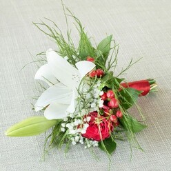 Captured in high-definition from above, this refined mini bouquet brings together the finest local floral traditions in Queensbury. The radiant centerpiece is a pristine white lily, unfurling in serene majesty as sunlight gently illuminates its arching petals, which are adorned with russet speckles near the core and vibrant yellow anthers-triggering a sense of pure anticipation, as if marking the start of a special ceremony along the Jubilee line. Tucked close to the lily, a striking red rose opens to reveal its velvet riches-a luxurious tribute to enduring love. Clusters of small, shining red hypericum berries nestle alongside, their festive gloss reflecting the energetic bustle of Queensbury's community celebrations. Wisps of baby's breath are scattered throughout, offering a soft silhouette against the dark green backdrop of ferns and leafy sprigs. A pale green lily bud extends to the left, promising blossoming joy for days to come. All the stems are expertly bound in a sleek red satin ribbon, adding both sophistication and a hint of theatrical flair. This compact bouquet, with its immersive reds, whites, and greens, and thoughtful local artistry, is ideal for wedding parties, anniversary gifts, or marking a momentous occasion within Queensbury's close-knit neighbourhoods-a refined floral gesture handcrafted for those who appreciate both beauty and meaning.