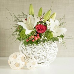 A refined floral centrepiece is styled as though ready to grace a wedding table in Queensbury, perhaps at an intimate reception just off Honeypot Lane. The arrangement is cradled in a spherical white vase with an intricate open-weave pattern, reminiscent of hand-woven wicker or delicate branches interlaced into a modern basket. Rising from this sculptural base is a lush display of white lilies, red roses, and textured greenery, carefully composed to balance drama with softness. The lilies are the stars: some fully opened with satin-smooth petals cupping golden stamens dusted with pollen, others still in tapered green buds that tilt gently upward, hinting at the scent that will unfold over the next few days. Tucked among them, rich red roses add depth and passion, with one particularly prominent rose in perfect spiralled form, its velvety petals layered tightly at the centre. Between these classic blooms sit bright green, globe-shaped pompons-likely dianthus or chrysanthemums-adding a playful, tactile element that feels fresh and contemporary, like a nod to Queensbury's mix of traditional and modern homes. Fine sprigs of white baby's breath float through the design, softening edges and giving an almost cloud-like lightness. Small, glossy red berries, reminiscent of hypericum, provide festive touches that would suit winter weddings or celebratory dinners. Long, slender fronds and deeper green leaves cascade gently from the arrangement, framing the flowers and enhancing their colour. Two pale, woven decorative spheres rest casually on the smooth white surface beside the vase, echoing its texture and bringing the entire scene together. A neutral, lightly textured background allows the crisp whites, romantic reds, and lively greens to stand out, evoking a sense of calm celebration and thoughtful craftsmanship by a skilled local florist.