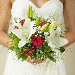 In the heart of Queensbury, a wedding morning is quietly captured as a bride holds a bouquet where every blossom tells a story. The snowy expanse of her strapless gown contrasts beautifully with the vibrant assembly in her hands: white oriental lilies opening in marvel, their wide petals and shining stamens catching the gentle morning light that streams near Queensbury Circle. Between these lilies nestle deep red roses, their velvety, spiraled petals evoking a quiet passion and timeless romance-a nod to long walks in Canons Park. Emerging green buds hint at future blossoms and new chapters, while baby's breath drifts like tiny, shimmering wishes among the larger blooms. Bright red hypericum berries offer joyful highlights, and playful spheres of green foliage bring an unexpected, tactile delight-adding depth reminiscent of nearby gardens in spring. Slender, feathery greens and broad leaves blend for a natural, hand-gathered finish, underscoring the bouquet's crafted freshness. It's more than a simple arrangement; it's a heartfelt celebration of commitment and beauty, the scents of lilies and roses mingling softly, destined to become part of cherished memories and the unique atmosphere of a Queensbury wedding.