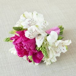 A small, exquisitely composed bouquet rests on a pale beige, linen-like cloth, the gentle weave of the fabric creating a calm, natural stage for the flowers to shine. At the centre is a bold fuchsia bloom, reminiscent of a garden peony at full flush, with tightly cupped, ruffled petals layered one over another to create a plush, cushiony dome of colour. Wrapped around this main flower are several crisp white alstroemeria, their long, slightly reflexed petals delicately painted with maroon and deep red streaks that radiate from their centres, adding fine detail like handwritten notes. The contrast between the intense magenta and the cool white petals gives the arrangement a sense of effortless summer elegance, as if captured between church and reception on a wedding day in Queensbury. Threaded through the design are slender, fresh green buds and foliage, upright and slightly spiky, which introduce movement and a hint of the garden, evoking the feel of stems just gathered from a local nursery or flower market. These greens gently frame the flowers without overpowering them, helping the eye travel from the vivid core to the softer edges. The stems are trimmed short and wrapped in a smooth, bright pink satin ribbon that mirrors the fuchsia bloom, its glossy surface hugging the stems and catching the light with a subtle gleam. The bouquet feels intimate, ideal for a groom's boutonnière, a bridesmaid's wrist corsage, or a keepsake posy for a special celebration. With its balanced mix of colour, texture, and thoughtful craftsmanship, it reflects the kind of romantic, modern floral work often requested from flower specialists around Queensbury.
