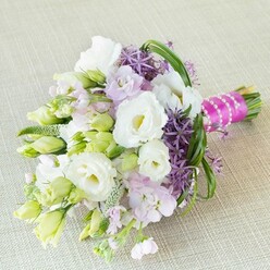Bathed in the gentle daylight so typical of a calm Queensbury morning, this exquisite bouquet rests across a light linen background, exuding an air of fresh optimism and understated romance. At its heart, creamy white Lisianthus flowers unfurl soft ruffled petals, some wide open to show their pale yellow centers, others still shyly held in tight, delicate buds-a nod to the anticipation surrounding a wedding in the heart of Queensbury. Pastel lavender-pink stock flowers nestle beside the Lisianthus, infusing the bouquet with a powdery sweetness evocative of spring strolls through Roe Green gardens. Vivid deep purple allium blooms, with their star-shaped florets fanning outward, deliver a modern, spirited contrast, hinting at the vibrant diversity found in local floral shops on Queensbury Circle. Interlacing the arrangement, slender, arching springs of fresh bear grass and a scattering of lime-green buds and filler create an organic, flowing silhouette that mimics the sweeping greens of the nearby parks. The bouquet's stems are expertly tied with a hot fuchsia ribbon, wrapped in an elegant line of glowing pearls that glint in the sunlight-details which elevate this from a mere arrangement to a treasured keepsake. Designed to celebrate unity, artistry, and memorable moments, this bouquet offers not only a feast for the eyes but a tactile sense of cool petals and crisp greenery. Whether destined for a city-centre ceremony or a local celebration, it stands as a radiant tribute to friendship, romance, and the craftsmanship found at the heart of Queensbury's floral community.