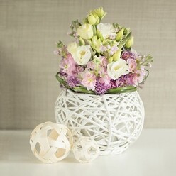 In a softly lit Queensbury dining room, a bespoke floral arrangement serves as a testament to the local florist's artistry and love for natural elegance. Creamy white and soft yellow eustoma unfurl in gentle waves, their ruffled petals recalling the delicate blooms found along the paths of Eton Grove Park. Pink stock (matthiola) flowers provide structural height, emerging from amongst the eustoma with a subtle pop of colour and the sweet aroma often cherished at early morning flower markets. Clusters of petite purple filler flowers settle at the base, adding depth and a playful vibrancy reflective of Queensbury's lively community gardens. Encircling the design, deep green foliage adds structure and frames the main display. All are gathered into a unique white, spherical vase, its open-work weave resembling modern sculptural art made from painted rattan or skillfully wrapped twigs. On the side, two decorative, basket-like rattan spheres-one slightly larger-offer a tactile, handcrafted element and reinforce the theme of local creative spirit. The entire composition is set against a softly textured, neutral-toned background that quietly amplifies the bouquet's gentle colours. This scene radiates an inviting, tranquil mood-perfectly suited for elevating wedding breakfasts, anniversary tables, or simply creating a welcoming atmosphere in any Queensbury home.