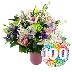 On a pure white backdrop, a luxurious celebratory bouquet radiates warmth from a sleek, ribbed, dusty-pink vase, the kind of arrangement a Queensbury florist might prepare for a once-in-a-lifetime 100th birthday along Honeypot Lane. The eye is first drawn to the luminous pink Oriental lilies, some wide open to reveal their long stamens, others still held in elegant buds that promise more beauty in the days ahead. Around them, soft blush roses and deeper dusty-rose blooms create a gentle gradient of colour, their velvety petals layered tightly near the centre and loosening at the edges like a well-loved heirloom book. Threaded between these classics, velvety purple lisianthus add richness and height, while sprays of small lavender asters, each with a golden centre, bring a sparkling, almost daisy-like lightness. Bright lime green button chrysanthemums peek through the petals like tiny fireworks, giving the design an upbeat, modern twist that feels right for a major milestone. Wisps of delicate white baby's breath float through the bouquet, softening transitions and lending a dreamy, cloud-like texture. A mix of dark and mid-green foliage cradles the flowers, with glossy leaves and slender stems framing the colours and grounding the whole display. Just to the lower right, a round foil balloon shines with a full-surface pattern of multicoloured confetti in vivid reds, oranges, yellows, greens, and blues, echoing the liveliness of local Queensbury celebrations. Bold white numerals "100" sit proudly at its centre, slightly raised in appearance, marking a century of memories. The overall impression is one of exuberant gratitude and festive elegance, ideal for same-day delivery in Queensbury to honour a beloved centenarian or a hundred-year anniversary.