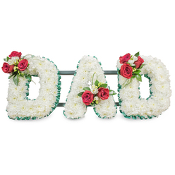 This meticulously made funeral tribute forms the word DAD in bold block letters, each letter entirely carpeted with countless small, immaculate white chrysanthemums that give the display a soft, cloud-like density and a feeling of serenity. A textured emerald-green ribbon outlines every letter, offering a crisp contrast that sharpens the typography and imparts a dignified frame to the white surface. Placed with considered balance, three floral clusters punctuate the composition: a rich spray of deep red roses and tiny white freesia on the upper curve of the left D, a matching cluster near the lower left diagonal of the central A where slender loops of green foliage lift the eye, and a mirrored arrangement on the upper curve of the right D. The roses' velvety red and the freesia's fresh white introduce warmth and scent against the chrysanthemum's clean coolness, while lush greenery adds depth and texture. Behind the letters, subtle dark green or black support rails are just visible, ensuring the large tribute remains steady when placed at a chapel or graveside. The lighting is even and soft, revealing petal detail without harsh shadow. Designed for memorial services around Queensbury - whether at a local chapel near Queensbury tube or a graveside in the borough - this tribute reads as a composed, heartfelt gesture of respect; chrysanthemums for devoted love and red roses for profound affection make it a timeless way to honour a father's memory.