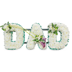 The image shows an elegant DAD funeral tribute, created as three bold, three-dimensional letters that stand side by side, all formed from masses of tightly arranged white chrysanthemums. Each tiny floret is fresh and full, combining to form a plush, cloud-like surface that suggests purity, peace, and a gentle final goodbye. Along the outer edge of every letter runs a rich, emerald green ribbon, slightly ruffled and satin-soft, which gives a clear outline and infuses the design with a touch of natural vibrancy. Behind the flowers, you can just make out slim dark bars that reveal a sturdy frame, allowing the piece to stand upright at a funeral service in Queensbury, perhaps at a chapel on Church Lane or in a quiet crematorium setting. The left-hand D carries a small, graceful posy of white freesia and lush foliage on its upper curve, the blossoms lifting the line of the letter with a subtle, hopeful air. Two slender green stems sweep diagonally from this first D towards the centre, connecting visually with the A and guiding the gaze along the tribute. Where these stems meet the base of the A, a deeper accent draws attention: two rich, purple calla lilies, with their smooth, sculpted trumpets, nestle among more white freesia and glossy greenery, creating a focal point that feels both modern and timeless. The final D is finished with a smaller cluster of freesia and foliage on its upper right, echoing the other side and bringing balance. Overall, it is a lovingly crafted memorial piece, ideal for a Queensbury family wishing to honour a father with dignity and heartfelt remembrance.