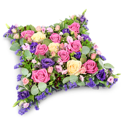 A meticulously crafted floral cushion, designed as a soft rectangular pillow, lies centred against a bright white background, giving every flower room to sing. The arrangement is densely woven with roses and seasonal blooms, creating a lush surface that looks almost like a quilt of petals. Deep fuchsia pink roses and softer pastel pink varieties are interlaced with creamy yellow and ivory roses, each bloom at its peak, petals gently unfurling to reveal intricate whorls and velvety texture. Nestled among them are rich, royal purple lisianthus, their layered, ruffled cups introducing a sophisticated depth of colour that anchors the design. Tiny clusters of lavender-toned filler flowers, such as statice or limonium, appear like delicate embroidery stitches, adding fine detail and a hint of wildflower charm. The entire composition is cradled in an abundance of foliage: round, silvery eucalyptus leaves and deeper green, fern-like fronds peep out between the flowers, bringing a cool, calming contrast and a whisper of woodland fragrance. The cushion's rounded corners and gently domed profile create a soothing, restful silhouette, suggestive of a head laid to rest in peace. On the white backdrop, a faint shadow beneath the arrangement gives a subtle sense of depth, as if the tribute has just been placed on a table in a quiet chapel or family home in Queensbury, perhaps near Kingsbury Road or one of the area's peaceful residential streets. This floral pillow radiates compassion and thoughtful care, a gentle, dignified way to honour a loved one and share unspoken feelings at a funeral or remembrance service.