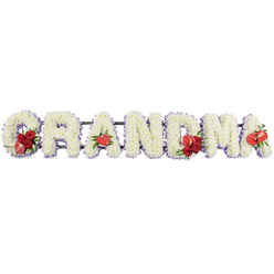 The image shows a beautifully composed funeral flower tribute spelling GRANDMA in large, upright capital letters, arranged in a straight line as if ready to be placed beside a coffin or at the front of a chapel in Queensbury. Each individual letter is built from tightly massed white chrysanthemums, their neat, button-like blooms pressed closely together to form a dense, cushiony surface with a soft, almost quilted appearance. Around the outside edge of every letter runs a slim band of pale lavender or light purple ribbon, giving the design gentle definition and adding a faint, soothing wash of colour that softens the starkness of the white. At key points along the lettering, rich red flowers introduce warmth and emotion: the G features a luxurious cluster of deep red roses combined with an eye-catching red and white variegated calla lily, tucked into the lower curve. The first A carries a single, shiny, heart-shaped red anthurium, positioned at its base like a small emblem of love. In the centre, the M is decorated with another group of red roses, surrounded by lush green foliage and partnered with a matching red anthurium. The final A is finished with its own solitary anthurium bloom, echoing the earlier accents and bringing balance to the piece. A dark green metal or plastic stand supports the letters just behind, keeping them aligned while remaining visually subtle. The tribute feels thoughtful and tender, the kind of bespoke arrangement a Queensbury family might choose from a trusted local florist to remember their grandmother with dignity and affection.