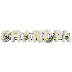 The image shows a beautifully composed funeral flower tribute spelling out GRANDPA in large, block capital letters, arranged in a steady horizontal line as though resting across the front of a coffin at a service in Queensbury. Each letter is built up from a dense covering of soft white chrysanthemums or carnations, every bloom set tightly together to create a smooth, cushion-like texture that feels gentle and forgiving. The surface of the letters is uniform and immaculate, with no gaps, reflecting the care and attention of an expert florist who understands how significant such a piece can be when a family gathers to say goodbye. Around the edges, a subtle outline of greenery and, in some places, a delicate edge of ribbon quietly frames the white flowers, giving extra definition to each character. Nestled within the expanse of white are thoughtfully positioned accents of deep purple and lilac-toned flowers, possibly lisianthus or similar seasonal stems, combined with glossy green foliage. These appear in the openings of the 'G', both 'A's, and the rounded part of the 'P', like gentle touches of colour symbolising cherished memories. The letters are supported by a dark green frame hidden beneath, ensuring that the tribute can be safely displayed at a chapel, church, or crematorium serving the Queensbury community. The overall feeling is one of quiet reverence and love, a personalised floral arrangement created to honour a grandfather's life with grace, suitable for same-day delivery to funeral homes and service venues across Queensbury and the surrounding streets.