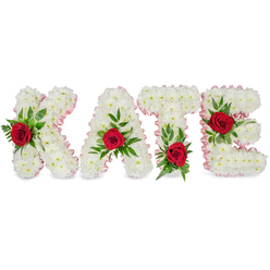 This photograph presents a bespoke floral name tribute reading KATE, crafted with the quiet precision of a seasoned florist and set against a clean white background so the arrangement's colours sing. Each of the four block letters is composed of tightly packed white chrysanthemums, their rounded blooms creating a plush, tactile surface that feels both serene and solid. Around the edges of every letter a soft pale pink satin ribbon has been carefully laid, the slight pleats and satin sheen adding a refined border that warms the otherwise pristine palette. A single, dramatic red rose punctuates the centre of each letter, its deep crimson petals contrasting vividly with the white chrysanthemums and serving as focal points of affection and remembrance. Lush green foliage - a mix of broad glossy leaves and delicate ferny sprigs - cradles each rose and adds sculptural depth and organic movement to the lettering. Placement is thoughtfully considered: the K's rose nestles on the lower part of its left vertical stem; the A's bloom sits at the tip of its apex; the T features its rose at the base of the left side of the horizontal crossbar; and the E bears its rose on the middle arm, each accent balancing the geometry of the type. The scent is quietly floral, a cool chrysanthemum base tempered by the warm, sweeter note of red rose. Ideal for a funeral or memorial service, this arrangement reflects care and craft familiar to florists serving Queensbury, nearby Harrow and the surrounding northwest London neighbourhoods.