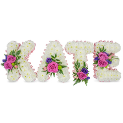 A tender, custom-made standing tribute spells KATE in bold, sculpted letters, each letter built from thousands of small, immaculate white chrysanthemums that form a continuous, cloud-like surface. The chrysanthemums are arranged so the letters sit three-dimensionally and feel plush to the touch, their tiny petals catching soft, diffused light that emphasizes the purity of the white. A delicate light-pink satin ribbon outlines each character, following inner and outer curves and imparting a faint, elegant blush that frames the white like a soft-edged photograph. Rich fuchsia-pink roses appear as focal notes, their velveteen petals layered and intimate, joined by pastel pink spray roses that lend a gentle sweetness. Cool, deep purple or blue accents - likely lisianthus or statice - thread through the design to deepen the palette and add quiet formality. Varied foliage, from lacy fern fronds to broad eucalyptus leaves, is woven around the clusters, introducing green relief, subtle scent, and natural movement. The floral groupings are carefully sited: a pronounced cluster along the K's diagonal stroke, a composed spray at the A's lower right curve, a base cluster on the T, and two composed accents on the E's centre and lower bar, creating balance and a reading that feels considered and personal. Set against a plain white studio background and presented upright as a memorial piece, this floral name tribute evokes solace, dignity, and the meticulous hand of a local Queensbury florist preparing a meaningful remembrance for a funeral or wake.