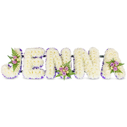 This deeply personal floral name tribute reads JENNA in substantial, three-dimensional capital letters crafted from small, snowy-white chrysanthemums that form a cushiony, tactile surface across each character. A subtle ribbon of purple or a rim of tiny lilac blooms outlines the inner edges of every letter, giving clarity and a soft halo of colour against the pristine white. Tasteful accent clusters punctuate the lettering: a compact spray of blush and lavender roses tucked into the sweeping curve of the J, another gathered at the lower centre of the second N, and a final grouping on the upper right of the A. Interspersed cream and pale-yellow roses add gentle tonal warmth while slim fern fronds weave fresh green texture through the clusters. The letters sit joined by slender dark support bars that create a secure, linear presentation suitable for placing at a chapel lectern or beside a memorial photograph. The arrangement exudes a hushed, comforting fragrance of roses and chrysanthemums and reads as both a visual nameplate and a heartfelt gesture. As an experienced Queensbury florist might suggest, this composition is designed to carry sentiment into local services at nearby community chapels, crematoriums, and family homes, offering a bespoke expression of remembrance that balances dignity with delicate floral craftsmanship.