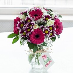 A richly coloured floral arrangement is showcased in a clear, jar-style glass vase, placed against a bright, softly blurred white background that feels like gentle daylight filtering through a Queensbury kitchen window. The bouquet is dominated by bold gerbera daisies in shades of deep fuchsia and magenta, their wide, layered petals encircling dark, velvety centres that give the design a confident focal point. Surrounding them are daisy-like chrysanthemums in striking purple and white bicolour, each bloom with delicately fringed petals that shift from intense plum at the tips to soft cream near the warm yellow centres, adding depth and intricate texture. Threaded among these statement flowers are clusters of creamy white spray roses, some opening to reveal their spiralled hearts, others still closed in tight buds that promise to unfurl over the coming days, lending a sense of gentle romance. Fine sprays of white gypsophila weave through the arrangement, creating a cloud-like lightness that softens the stronger colours and echoes the look of spring blossoms in Queensbury Park. Dark, glossy greenery cradles the flowers, adding structure and contrast, while also suggesting the freshness of a bouquet prepared early that morning. The stems are visible through the glass, neatly trimmed and fanned out in the water, and the vase's neck is wrapped with a simple length of rustic twine tied into a modest bow. From this bow hangs a small, rectangular white gift tag featuring a delicate pink rose motif and subtle text, hinting at a personalised message from sender to recipient. The overall mood is joyful yet refined, perfect for marking birthdays, congratulations, or just-because deliveries to homes and workplaces across Queensbury, capturing the uplifting feeling of receiving flowers on an ordinary day.