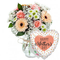 Set against a clean white backdrop, this Mother's Day ensemble looks like a moment of celebration paused in time, ready to be carried to a doorstep in Queensbury. The focus is a generous mixed bouquet arranged in a clear glass vase, its jar-style shape simple and honest, with a neat band of rustic twine tied around the neck to suggest a florist's careful hands. The flowers themselves form a soft dome of colour: peach-pink roses fully open, their layered petals curling outward in perfect spirals; bold peach gerbera daisies with dark chocolate centres offering a vivid contrast; and daisy-like white chrysanthemums with fresh apple-green centres peeking between the larger blooms. Floating through the arrangement, clouds of white gypsophila add an airy, almost cloud-like lightness, while rich green foliage creates depth and frames the pastel tones with a natural, gardeny feel. Tucked modestly behind the flowers is a small white card, only a corner visible, suggesting a hidden handwritten message from child to mum. In front, leaning toward the viewer, is a large heart-shaped foil balloon tethered to the vase. Its bright white surface is sprinkled with shiny gold and silver confetti dots, catching the light like tiny sparks of joy. In the middle of the balloon, a soft peach heart holds the phrase "HAPPY Mother's DAY" in bold black letters, the word "Mother's" rendered in a more flowing script that feels tender and personal. The composition as a whole conveys freshness, affection, and the sort of thoughtful surprise you might send across Queensbury for same-day Mother's Day delivery.