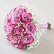 Set upon a softly textured, cream-hued background, this exquisite round bouquet brings together the very best of local floral artistry near Queensbury. The lush blossoms of about ten to twelve roses take centre stage, their inner petals a powdery lavender-pink, fading gracefully into fuchsia along the tips, forming a layered visual depth treasured by any romantic at heart. Interlaced with these blooms are generous clouds of pure white baby's breath-tiny, starry flowers that lighten the bouquet and evoke a sense of gentle, airy elegance perfect for wedding mornings in the area. The well-kept stems, fresh and still tinged a lively green-brown, are wrapped tidy with a sheen of pink satin ribbon, its elegant spiral completed by a string of lustrous little pearls. As sunlight filters gently across the bouquet, the textures-from velvety petals to the feathery gypsophila-are softly illuminated, casting subtle shadows that add to its allure. This arrangement from a Queensbury florist, crafted with care and a keen sense of tradition, makes a sophisticated gift or a heartfelt finishing touch for bridal celebrations, anniversaries, or memorable moments shared in the warm embrace of this local community.