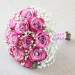 A beautifully composed bouquet of pink-lavender roses and airy baby's breath lies at a gentle angle across a soft, light beige fabric backdrop, the neutral surface echoing the calm skies often seen over Queensbury Park on a summer morning. At the centre, about a dozen lush roses cluster together in a compact dome, their petals transitioning from muted mauve and lavender-pink within to vibrant raspberry, fuchsia, and deep magenta at the edges. Each flower is either tightly furled or just beginning to unfurl, revealing the complex swirl of layers that give garden roses their romantic reputation. Fine sprays of white Gypsophila surround and thread between the roses, forming a delicate halo that lightens the overall look and adds a misty, almost ethereal quality. The baby's breath appears like tiny pearls of light, balancing the strong rose colours and creating a sense of gentle movement around the main blooms. A few hints of rich green foliage peek out near the base, reminding you that these are freshly cut stems, likely prepared that morning in a local Queensbury florist studio. The stems themselves are neatly bound at the lower right, cocooned in a smooth, lilac-toned satin ribbon whose soft sheen catches the light. Around this ribbon, a fine strand of white pearls spirals gracefully, suggesting bridal tradition and careful craftsmanship. The photograph captures a bouquet that feels made for a wedding aisle, a civil ceremony at a nearby registry office, or an intimate vow renewal, offering a refined expression of love and tenderness for couples celebrating their story in and around Queensbury.