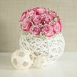 A serene indoor scene shows a rounded bouquet of soft pink roses and delicate white baby's breath nestled into a white, woven spherical vase, arranged as though it's just been set down on a bright windowsill in a Queensbury home. Around twenty roses form a lush dome, each bloom opening in layers from a pale, almost blush centre to richer pink and near-fuchsia edges on the outer petals. Between the roses, tiny stars of Gypsophila float like clouds, softening the shape and giving the arrangement a romantic lightness. The flowers sit in a distinctive globe-shaped container crafted from interlaced white strands that resemble painted wicker or rattan; its open, basket-like pattern lets light pass through and creates gentle shadows on the smooth white surface below. To the left of the vase, two decorative orbs rest side by side, each fashioned from thin, natural wood or fibre strips in a loosely woven, geometric pattern, their warm beige tones echoing the subtle cream in the background wall. The wall itself is a warm, lightly textured beige, similar to what you might find in a cosy living room off Queensbury Circle, adding calm and allowing the pinks and whites to stand out clearly. Soft, even lighting illuminates the scene without harsh contrast, catching the curve of each petal and the fine threads of the woven decor. The overall mood is peaceful and refined, like a thoughtfully chosen centerpiece for a quiet anniversary dinner or a Mother's Day surprise, the kind of handcrafted arrangement locals might choose for intimate gatherings or small celebrations at home across Queensbury.