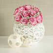 A rounded bouquet of candy-pink roses is perched gracefully on a woven white globe, creating a modern wedding piece that balances romance with architectural style. The roses form a smooth dome, petals tightly layered in spirals that move from pale, almost porcelain pink at their hearts to deeper magenta along the edges, as though each bloom has been gently kissed by evening light. Wisps of white gypsophila, or baby's breath, nestle around the base of the cluster, their tiny, cloud-like blossoms giving the roses a soft, dreamy frame. The arrangement rises from a striking white orb vessel made of hand-woven strands that crisscross and curve, leaving airy gaps where light passes through and dances on the tabletop. From some angles, the globe appears almost like a piece of modern sculpture, its smooth, matte finish contrasting with the plush texture of the flowers above. Two small geometric ornaments sit at the foot of the orb, enhancing the sense of contemporary styling without stealing focus. The whole composition is set against a bright, uncluttered background, as if placed near a sunlit window in a Queensbury townhouse close to Queensbury Station or along Kenton Road. You can almost imagine the gentle scent of roses drifting through a reception venue, calming and welcoming guests. This design feels perfectly at home at minimalist weddings, intimate engagement parties, or as a statement piece in a stylish Queensbury living room, offering a quiet yet confident celebration of love and modern elegance.