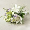 Resting gracefully on a subtly woven, pale linen fabric, this bridal bouquet brims with the classic elegance and freshness that define Queensbury's wedding celebrations. The star attraction is an array of large, trumpet-shaped white Oriental lilies, their pristine blooms open wide or just beginning to unfurl, each revealing intricate speckling and vibrant golden anthers at their centres. Mottled between these lilies are two-toned lisianthus, their delicate white petals trimmed in bold purple, blending seamlessly with clusters of smaller, rich purple Veronica spikes that reach upward with lively charm. Wisps of soft white gypsophila peek through the arrangement, providing gentle, cloud-like volume and contrast to the saturated hues. Surrounding the flowers is a lush medley of greenery-broad, deep leaves provide a steady base, while feathery asparagus fern trails outward, imparting a fresh sense of movement. The bouquet's stems are bound with a pure white ribbon and displayed horizontally so the artistry of each blossom is fully revealed, reminiscent of bouquets prepared with loving care by Queensbury's talented local florists. Ideal for a wedding in the area, an anniversary, or as a sophisticated gift, this arrangement brings together purity, sweet fragrance, and a touch of modern romance, suitable for every special moment across Queensbury.