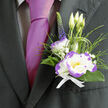 Nestled against the deep charcoal fabric of a well-cut suit, this boutonnière speaks of considered craft and gentle confidence familiar to anyone ordering locally from Flower Delivery Queensbury. Its focal bloom is a white lisianthus with violet-purple edging-the petal tips blush with colour so that each fold looks hand-painted. Surrounding the main flower, tiny clouds of gypsophila lend a whispered innocence, while slender, looping blades of ornamental grass bring a soft, kinetic texture as if the piece might stir with a light breath. Tall, unopened lisianthus buds introduce a sense of possibility, and a narrow spike of indigo veronica provides a refined vertical accent that keeps the eye moving. Dark green foliage frames the composition, glossy where the light catches it, matte where it recesses, creating depth against the suit lapel. Everything is bound with a crisp white ribbon whose smoothness contrasts pleasantly with the natural textures of stem and leaf. The juxtaposition of the boutonnière with a magenta silk tie gives the ensemble a contemporary edge-perfect for a groom stepping out near Queensbury station or a guest arriving for a milestone celebration on the high street. The scent is barely there: a clean, green whisper that suggests newly cut stems and early-market blooms. As a local florist I design pieces like this to feel both sophisticated and personal, reliable companions for long days of celebration and small moments you want remembered.