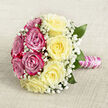 Set against a neutral, woven background that looks like soft oatmeal-coloured linen, this hand-tied bouquet glows with the romance and care of a carefully planned Queensbury celebration. The flowers are arranged in a rounded, almost perfectly circular form, divided into two harmonious colour fields: one side is filled with plush fuchsia and magenta-pink roses, their velvety petals spiralling outward in generous layers; the other half features equally full roses in a gentle, buttery cream with the faintest suggestion of lemon at the centre. Sprays of delicate white baby's breath weave between both tones, forming a light, airy mist that softens the transition and adds a traditional bridal elegance. Subtle, deep green foliage peeks between the blossoms, giving depth and contrast, like small notes of the parks and gardens tucked around Queensbury. The stems are expertly bundled into a clean hand-tied handle, wrapped tightly in lustrous pink satin ribbon that catches the light with each fold. Around this, a string of small white pearls curls in a precise spiral, echoing the curves of the rose petals and adding a touch of luxury often chosen for weddings and engagement parties near Queensbury Station or along Honeypot Lane. The bouquet appears freshly made, with no bruising on petals or leaves, and you can almost imagine a soft rose fragrance rising from it. The overall mood is calm, joyful, and intimate, as though this arrangement is waiting to be lifted by a bride, bridesmaid, or partner about to surprise someone special with a declaration of love.