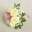 A petite, hand-crafted bouquet is presented on a diagonal angle against a pale beige, finely textured fabric backdrop that looks like smooth linen, giving the whole scene a calm, understated elegance. At the centre, five creamy ivory roses bloom with soft, layered petals that curl outward in gentle waves, their tone warm and inviting, like candlelight on a late afternoon in Queensbury. Just to the front-left, a single fuchsia rose stands out boldly, its saturated pink petals offering a lively contrast to the softer white tones and instantly becoming the focal point of the arrangement. Around and between the roses, generous tufts of white baby's breath create a misty, cloud-like effect, their tiny blossoms adding both volume and a delicate, romantic air. Deep green leaves with pointed tips provide a crisp frame, the foliage grounding the bouquet and adding depth, as if freshly gathered from a well-tended garden near Queensbury Park. The stems are tightly wrapped in a bright pink satin ribbon, its glossy surface smooth and luxurious to the touch, shaped into a neat handle. Across this ribbon, multiple horizontal rows of small, iridescent pearls are meticulously placed, catching hints of light like dew drops and elevating the bouquet into something clearly meant for a special moment. The close-up, slightly elevated view allows every flourish to be appreciated, from the texture of the fabric beneath to the precise positioning of each bloom. It feels perfectly suited to a wedding corsage, boutonnière, or small bridesmaid's bouquet arranged for a ceremony near Queensbury station or a family celebration at home.