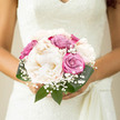 This image captures a romantic bridal bouquet resting lightly against what could be the folds of a wedding dress, a serene combination of creamy peonies, pastel roses, and gypsophila arranged with the care of an experienced Queensbury florist. At the centre, the peonies are the main focus: full, rounded blossoms in soft cream and off-white, each petal slightly ruffled at the edge, giving a plush, cushiony impression that feels both luxurious and gentle. Tucked between them are roses in dusty pink and muted lavender, their velvety petals spiralling outwards in graceful layers, bringing depth of colour without overpowering the calm palette. Wisps of baby's breath weave delicately through the blooms, its tiny white flowers scattered like confetti, evoking that fleeting moment when newlyweds step out into the daylight after saying their vows. Glossy green foliage encircles the bouquet, the broad leaves providing a defined outline and a fresh contrast to the soft pastel centre, grounding the design visually and adding a sense of natural lushness. The bouquet appears hand-tied, compact yet airy, the flowers drawn close together yet given enough room to show off their individual shapes and textures. The lighting feels soft and natural, as though the photo were taken near a window overlooking a quiet Queensbury street, lending the arrangement a tranquil glow. The overall effect is one of understated elegance and calm happiness, perfectly suited to a refined wedding at a local registry office or a nearby garden venue, where every detail is chosen to echo harmony, grace, and heartfelt love.
