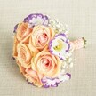 A radiant floral bouquet exuding romance and sophistication is presented from a high vantage point, gracefully resting atop a soft, oatmeal-hued textured fabric-an image that invokes the welcoming ambiance of Queensbury's local gatherings. At the core of the arrangement are lush, open peach and apricot roses, their velvety petals revealing gentle blush and creamy tones that seem to come alive in the soft natural light. Encircling these, refined Lisianthus blooms shimmer in crisp white, their frilled petals each traced with artistic streaks of deep purple and violet, adding a whimsical counterpoint to the warm rose palette. Thin clouds of Baby's Breath drift effortlessly among the flowers, imparting a weightless, classic charm reminiscent of traditional wedding scenes near Canons Park. A peach-pink satin ribbon, expertly wrapped and adorned with dainty pearl beads, not only secures the neat stems but adds an understated sense of occasion-crafted for hands that will carry it into momentous events. Designed with the local spirit in mind, this bouquet reflects the elegance and joy of Queensbury celebrations, whether as a bridesmaid's bouquet, an engagement centrepiece, or a heartfelt gesture for those who cherish exquisite blossoms and artisanal detail in North West London's vibrant community.