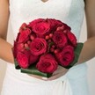 In this Queensbury-inspired wedding scene, a lush bridal bouquet emerges as a poetic centrepiece-velvety deep red roses, some unfolding in a spiral of soft petals, others remaining secret in tight buds, signal devotion and enduring love. Dotted among them, vivid clusters of red hypericum berries catch the light, their gloss reminiscent of fresh fruit from local Queensbury produce stands, adding dimension and playful detail. The flowers are impeccably gathered into a rounded, compact form and secured with broad arcs of glossy green foliage, creating a sculpted botanical collar that elegantly supports the arrangement. The bouquet rests gracefully in the bride's hands, her nails polished and her delicate touch suggesting anticipation and joy. The white wedding dress, its bodice tenderly adorned with lacework, frames the bouquet's intensity, letting rich reds and greens stand out against soft neutral tones in the background. The entire scene-bright, focused, and inviting-feels like a snapshot of love's fullness on a special day near Roe Green Park, blending personal celebration with community spirit. This bouquet is a sensory testament to joy, unity, and the craftsmanship of Queensbury florists, perfect for couples wishing to carry a piece of their neighbourhood's heart into married life.