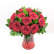 A classic, full bouquet of deep red roses stands proudly against a pure white studio background, every bloom turned forward as if offering a heartfelt message. Around a dozen or more roses, rich as burgundy velvet, are gathered in a generous, domed cluster, their petals spiralling inward to tight, romantic centres. The flowers are at perfect maturity, with no sign of wilting, so each head appears plush and luxurious, almost touchably soft. Between the roses, dark, glossy green foliage, likely Ruscus, slips in and out of view, its elongated leaves framing the blossoms and giving the arrangement a natural, garden-fresh fullness. The greenery provides a cool contrast to the passionate red, helping to define the rounded outline of the bouquet and softening its edges. All of this is cradled in a translucent ruby-red glass vase with a gently flared rim and base, the surface subtly ridged or swirled so that the light catches and shimmers across it. Through the tinted glass, the straight, dark stems are clearly visible, crossing and overlapping to create vertical lines that anchor the composition. The bright, even lighting has a daylight clarity, highlighting every curve of petal and sheen of leaf without casting harsh shadows, making the colours feel true and intense. This arrangement feels like an unspoken declaration of love or devotion, ideal for celebrating anniversaries, Valentine's Day, or a bold romantic gesture delivered to a home just off High Street in Queensbury, a cosy flat near the station, or a favourite restaurant for a surprise evening together.