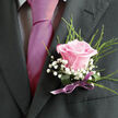 A romantic boutonniere featuring a single, perfectly formed pale pink rose is shown resting against the lapel of a tailored dark suit, the subtle lighting highlighting every curve of the bloom. The rose itself has that velvety, just-open quality that florists love to work with, its petals unfurling in a spiral of soft blush tones that deepen slightly toward the centre, suggesting the quiet excitement of a wedding day in Queensbury. Around the rose, fine stems of white gypsophila, or baby's breath, create a halo of tiny star-like blossoms, their delicate clusters echoing the lightness of confetti drifting outside local venues near Honeypot Lane. A few sprigs of airy greenery arch gracefully outward, adding movement and a fresh, garden-like feel, the greens providing a gentle contrast to both the blush rose and the rich charcoal fabric of the suit. The stems are neatly bound at the base with a narrow satin ribbon in a muted lilac or soft mauve, its subtle shine catching the eye without overpowering the flowers, reminiscent of the sheen of polished shoes or a silk tie worn to a celebration. The composition is intimate and refined, the kind of carefully crafted piece chosen for grooms, best men, or prom dates heading to events around Queensbury and neighbouring areas. Every element speaks of attention to detail-the balanced proportions, the mix of textures, and the harmony of colours-creating a small, wearable arrangement designed to look elegant in photographs and feel meaningful in the moment.