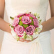 This bridal bouquet rests against the intricate lace of a wedding gown, capturing a moment of stillness before the celebrations begin in Queensbury. The eye is first drawn to the lush roses at the centre, their colour a refined blend of lavender, lilac, and dusty pink that feels both contemporary and timeless. Each rose is fully formed yet not over-open, with layers of petals curling softly outward like small, contented sighs. Tucked between them, lime-green button chrysanthemums peek out, their rounded, textured blooms adding a playful spark of colour and a hint of modern design. Around the outer edge, creamy white flowers and slender buds create a soft halo, almost cloud-like, gently framing the richer tones while keeping the overall palette light and bridal.
The bouquet is round, hand-tied, and neatly compact, sitting snugly in the bride's hands as she prepares to walk down the aisle, perhaps at a nearby church or venue off Honeypot Lane. The stems are trimmed evenly, likely wrapped in satin or ribbon that disappears into the folds of the dress, giving the impression that the flowers simply float in front of her. Soft, natural light washes over the arrangement, highlighting the velvety texture of the rose petals and the almost dewy freshness of the green chrysanthemums. You can almost imagine the scent: the calm, familiar perfume of garden roses mingling with fresh greenery, soothing and uplifting at once. Created with the experienced eye of a local Queensbury florist, the bouquet is designed not only to look beautiful in the first photos, but to carry its colour, form, and gentle joy right through to the last dance of the evening.