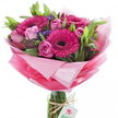 A joyful hand-tied bouquet sits upright in a clear, cylindrical glass vase, its bundled stems visible through the water and glass like the spine of a living painting. The composition is anchored by several large gerbera daisies in a vivid fuchsia-pink, each with a deep, almost black centre that draws the eye and gives the arrangement a confident, dramatic focus. Nestled around those bold blooms are soft pink roses in a variety of stages - from tightly coiled buds that promise unfolding fragrance to fully open roses showing layered, satiny petals - their pastel tones calming the intensity of the gerberas. Lush green foliage, a mix of broad, smooth leaves and more slender stems, forms a verdant scaffolding that lifts and separates the flowers, while small clusters of deep purple-blue filler - likely statice - thread through the bouquet to add fine texture and a cool counterpoint to the warm pinks.
The flowers are wrapped in crinkled, sheer pink cellophane that graduates from a pale, almost translucent halo at the top to a richer, more saturated pink toward the base, creating a soft, celebratory bloom of colour around the heads of the flowers. A length of rustic twine is tied around the neck of the vase, from which hangs a small white rectangular tag printed with a stylised pink-and-green rose logo, a quiet signature of craft and care. In gentle morning light the glossy surfaces catch highlights and the scent reads as softly sweet and green - a bouquet that radiates uplift and bright celebration, perfect for birthdays, new beginnings, or simply to bring cheer to someone in Queensbury.