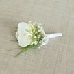 A refined orchid boutonnière is presented on a softly woven, light beige fabric, evoking the feel of a tailored suit laid out in a Queensbury home just before a wedding ceremony. At the centre, a single white orchid bloom opens with effortless grace, its velvety petals broad and unblemished, curving gently outward to create a rounded, sculptural form. At the heart of the flower, a hint of warm cream and soft yellow glows within the throat, introducing a quiet depth that keeps the white from feeling stark, more like candlelight than bright glare. Framing the orchid, clusters of delicate baby's breath scatter in all directions, their countless miniature white flowers forming a gauzy, cloud-like halo that softens the strong silhouette of the central bloom. Threaded beneath and slightly beyond these blossoms, fine strands of vivid green foliage arc and taper, adding fresh colour and a sense of movement, like a whisper of breeze coming up from the direction of Queensbury Circle. The stems have been meticulously trimmed and aligned, then cloaked in smooth white satin ribbon, wrapped snugly to create a slender, comfortable handle that would sit securely on a lapel during hours of celebration. Near the top, a neat satin bow, handcrafted and symmetrical, adds a classic detail that suggests care and craftsmanship. The light tan, linen-like fabric beneath shows a delicate weave, providing a softly textured, neutral stage that allows every nuance of petal, leaf, and ribbon to stand out without distraction. The overall mood is one of calm sophistication and heartfelt romance, the kind of orchid buttonhole a groom might choose for a church wedding near Queensbury, where elegance is expressed through purity, balance, and restraint.