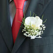 A refined orchid boutonnière rests neatly in the pocket of a dark, tailored suit jacket, capturing the mood of a polished celebration in Queensbury. At its heart, a single white orchid opens in full bloom, its petals creamy and luminous, curving outward with a quiet elegance that suggests purity and commitment. The orchid's centre holds a subtle touch of warm yellow or gold, like a hidden spark of emotion at the core of an otherwise calm design. Encircling the bloom, clusters of baby's breath float in soft, cloud-like formations, each tiny white flower adding a layer of delicacy and romance. Fine green foliage weaves in and out of the composition, bringing movement and life, as if the florist had just gathered fresh stems from a local flower stall near Queensbury's shops. A narrow white ribbon wraps the base, its tidy bow revealing the careful craftsmanship behind this seemingly simple piece. The boutonnière is framed by the structured lines of a charcoal suit and a striking red tie, whose strong colour provides a vivid backdrop that makes the white florals appear even more pristine. The overall effect is one of balance: modern yet traditional, refined yet heartfelt, suited to a wedding day walk from home to a nearby Queensbury ceremony, or a special event shared with family and friends. Every texture, from the orchid's velvet softness to the feathery baby's breath, invites an almost tactile appreciation, as though you could reach out and feel the cool petals and fresh greenery against your fingertips.