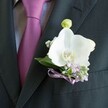 A close-up photograph shows a sharply tailored charcoal suit jacket, its left lapel in crisp focus, carrying an elegant white orchid boutonniere that feels perfectly suited to a wedding morning in Queensbury. A finely woven jacket in deep charcoal grey borders the scene, with a light grey dress shirt peeking through, patterned with the faintest texture. Against it rests a rich magenta tie, almost plum in tone, tied with smooth precision and giving a subtle sheen whenever the light catches its folds, reminiscent of late-afternoon sun over Queensbury Park. Pinned neatly to the lapel, the Phalaenopsis orchid dominates the composition: broad, velvety petals open in a soft, creamy white, curving gently around a vivid centre of yellow and warm orange. Around the orchid, the florist has tucked tiny clusters of baby's breath and delicate lilac blossoms, their pale lavender tones softening the contrast with the dark jacket. Slender sprigs of greenery and a single, pale green orchid bud stretch upward, suggesting fresh growth and the quiet anticipation of the ceremony. At the base, a narrow lavender satin ribbon is tied into an intricate little bow, its smooth, light-reflecting surface echoing the nearby tie and hinting at thoughtful colour coordination. The overall mood is calm, romantic, and meticulously composed, as if the groom has just stepped out of a car on a tree-lined Queensbury street. You can almost imagine a faint, clean floral scent rising from the orchid, mingling with crisp suit fabric and cool morning air, marking this as a moment of commitment, grace, and understated style on a special local wedding day.