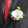 A close-up photograph focuses on a single wedding boutonniere pinned precisely to the lapel of a dark charcoal grey suit jacket, the fine woven texture of the fabric clearly visible beneath the flowers. At the heart of the design, a large white Phalaenopsis orchid opens its velvety petals, creamy and luminous, with a distinct yellow-and-white lip curving gently from the centre. Just above, two small, rounded lime-green button chrysanthemum blooms nestle into the composition, their tight, textured heads adding a lively contrast against the smooth orchid. Tiny clusters of white baby's breath float around the main flowers like a soft cloud, their delicate blossoms catching the light and softening the edges. Behind everything, a few rich green leaves create a natural backdrop, their sheen echoing the freshness of flowers just prepared by a careful florist. The base of the boutonniere is bound neatly with a narrow deep red satin ribbon that subtly mirrors the bold, solid red satin tie lying to the left, set against a finely checked pale grey and white dress shirt. Light falls across the arrangement in a gentle wash, highlighting the orchid's contours, the crispness of the shirt, and the refined structure of the suit's lapel and pocket seam. The overall impression is of quiet sophistication and romantic purpose, the kind of thoughtful finishing touch you might see on a groom heading to a ceremony at a Queensbury registry office or a nearby church. It feels like a small, wearable keepsake of the day-a calm, elegant accent designed to accompany vows, photographs, and happy celebrations with family gathered from across Queensbury's tree-lined streets and local parks.