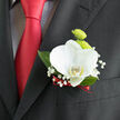 Pinned neatly to the lapel of a tailored charcoal suit, this orchid boutonnière offers the kind of quiet elegance that suits a wedding at a Queensbury registry office or an intimate reception off Kenton Road. At its heart, a flawless white orchid opens like a calm, sculpted shell, each petal broad and velvety with a soft yellow blush at the centre that draws the eye inward. Behind the orchid, two compact lime-green button chrysanthemums sit tightly together, their domed, cushiony heads creating a playful contrast to the orchid's smooth expanses of white. Finer details unfold in a halo of baby's breath that circles the focal blooms, clusters of minute, cloud-like flowers giving a sense of movement and lightness, as if a gentle breeze had passed across the arrangement. Deep, glossy leaves tuck in behind, grounding the composition with a rich forest green that feels stable and reassuring. At the base, a slim ribbon of deep red is wrapped with precision, its tone echoing the polished red tie that drapes down the front of the suit, tying floral and fabric together in one cohesive gesture. The lighting is soft and flattering, allowing the textures-the orchid's satin-like surface, the chrysanthemums' delicate ridges, the ribbon's subtle sheen-to stand out without harsh shadow. Crafted by an experienced local florist, the boutonnière has been assembled with care so it sits close to the chest, secure yet almost weightless, offering a fresh, contemporary nod to tradition for anyone celebrating love and commitment around Queensbury.