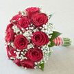 A romantic bouquet of red roses and baby's breath is shown in soft daylight, as if resting on a polished surface in a quiet Queensbury living room, moments before being handed to someone special. Each rose is a lush, velvety crimson, perfectly opened to reveal spiralled layers of petals that suggest both richness and warmth. Nestled between them, tiny white gypsophila blossoms cluster like a fine mist, their airy form giving the arrangement a graceful, cloud-like halo. This interplay of strong red and delicate white creates a sense of balance, where intensity and gentleness live side by side. Slender green leaves frame the blooms, adding structure and a hint of the garden, while the stems are gathered neatly into a hand-tied posy. The handle is wrapped in smooth, elegant ribbon, detailed with a line of small pearls that catch the light and add a subtle note of ceremony, echoing the refined style of wedding bouquets often seen leaving nearby Queensbury churches and registry offices. The bouquet feels at once classic and modern, as suitable for a bridal aisle as for an intimate proposal dinner at home. Imagining the scene, you can almost detect the soft, sweet scent of the roses blending with the barely-there freshness of the gypsophila, the overall impression one of quiet celebration and heartfelt devotion, carefully crafted by a florist who understands how flowers speak when words fall short.