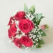 Swept up in the gentle spirit of Queensbury, this hand-tied bouquet unfurls a scene of timeless romance and local craftsmanship. At its core, five to six vibrant, velvety red roses blossom fully, each petal arranged in a classic spiral and revealing shades of rich crimson that conjure feelings of passion and celebration. Inviting the eye and heart alike, countless tiny clouds of airy white baby's breath (Gypsophila) weave between the roses, casting a soft halo of lightness against the bold backdrop of red. Lush, verdant green foliage-perhaps polished ruscus or fresh salal-clusters around the blooms, granting texture and life to the arrangement's profile. At the bouquet's base, sturdy stems are brought together and meticulously bound with an eye-catching red and white striped ribbon, which is delicately adorned with a trailing string of small, gleaming white pearls, suggesting both glamour and loving attention to detail. Resting upon a pale cream or off-white textured fabric-evoking the comfort of a familiar Queensbury home or a sunlit wedding venue-each element is perfectly highlighted under soft, even lighting. This bouquet is more than a floral arrangement; it is a statement, perfectly suited for weddings near Roe Green Park, anniversary celebrations, or as a heartfelt romantic gesture. Its subtle fragrance of rose and greenery brings a gentle, comforting air, infusing any special occasion in Queensbury with warmth, elegance, and an enduring sense of beauty.