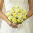 Held close to the bodice of a bridal gown, this bouquet of pale yellow roses reads as an intimate study in warmth and restraint-the kind of arrangement we craft at Flower Delivery Queensbury for ceremonies across Queensbury and nearby Fryent Country Park. Each rose is a soft butter-yellow, petals unfurling in smooth spirals to form immaculate, cup-like blooms that reflect the soft, diffused light. Wisps of white baby's breath are woven through the roses, their tiny, cloudlike heads creating a delicate veil that seems to float above the cluster and bring an airy luminosity to the palette. Encircling the composition are curved, ribboning blades of deep green grass that give the bouquet a refined, almost architectural border, balancing the floral softness with clean, botanical lines. The scent is mild and fresh-a light honeyed sweetness tempered by a green, dewy note-so unobtrusive it feels like a comforting memory rather than a headline. The arrangement is hand-tied and compact, made to sit comfortably in the bride's hands as she moves; the finish is neat and considered, the stems held together to form a gentle dome. Stylistically the bouquet whispers rather than shouts: romantic and hopeful, it is an emblem of steady affection and new beginnings. Whether photographed against the lace of a vintage gown or carried outdoors for portraits by local green spaces near Queensbury station, this bouquet brings a sense of serenity and refined craftsmanship to the wedding moment.