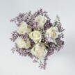 A high-angle overhead photograph reveals a compact, hand-tied bouquet arranged in a perfect circle, as if cradled gently in two hands. At the heart of the design sit six creamy white roses, each bloom impeccably formed, with tightly layered petals that spiral from a soft, cupped centre out to smooth, rounded edges. Their ivory-white colour carries the faintest whisper of green at the base of a few outer petals, a natural detail that adds freshness rather than flaw. Around these serene roses, a halo of tiny lavender and muted lilac statice blossoms forms a soft, cloud-like ring. The filler flowers create a fine, feathery texture, with many delicate stems and clusters that look almost like a haze of purple mist enveloping the roses. The lighting is gentle and even, as though the bouquet has been placed near a window on a calm day in Queensbury, with no harsh shadows to distract from the flowers' textures and tones. The arrangement rests on a clean, pale surface-off-white or light grey-reminiscent of a simple tabletop in a quiet home off Deansbury Drive. This neutral backdrop allows the white roses and lilac statice to stand out as the sole focus. The overall effect is one of calm sophistication and quiet romance, suitable for an intimate wedding, a sympathy gesture, or a thoughtful everyday delivery in Queensbury. Viewed from above, the bouquet feels complete and harmonious, a soothing, refined floral moment captured in a single serene frame.