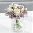 A close-up photograph captures a tranquil, elegantly styled bouquet where creamy white roses and clouds of lavender-hued statice are the gentle heart of the design. Each rose sits perfectly formed, its velvety petals opening in soft spirals, with the faintest wash of pale butter-yellow at the centre that hints at early-morning freshness. Around them, the statice stretches in airy clusters of tiny, papery blooms, their lilac and lavender tones creating a mist-like halo that softens the whole arrangement. The flowers are gathered into a clear glass vase with a distinctive crackle, or ice-effect, texture; the surface shimmers subtly as it catches the natural light, almost like frost on a Queensbury windowpane in early spring. Through the clear water, the straight green stems are visible, neatly trimmed and thoughtfully arranged, adding structure and a sense of natural calm. The bouquet rests on a flawless, bright white tabletop that reflects the light and casts soft, feathery shadows under the vase, while the background is gently blurred to a pale grey, keeping every bit of attention on the flowers. The atmosphere feels quiet and restorative, like a peaceful corner of a living room near Queensbury Park on a bright but still day. This is the kind of understated arrangement you might choose to soothe a hospital bedside, send a gentle condolence, or bring a serene touch to a home office in NW9, its delicate tones and refined composition speaking in a low, comforting voice rather than a shout of colour. The image honours the simple pleasure of fresh roses and statice, meticulously arranged by a skilled local florist who understands how to balance purity, texture, and light.