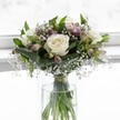 An elegant bouquet of white roses and seasonal blooms stands in a simple clear glass vase, softly illuminated as if placed on a windowsill in a quiet Queensbury home. At the centre, three full, creamy white roses open in perfect spirals, their velvety petals folding gently back to reveal warm ivory hearts that suggest innocence, grace, and freshly cut luxury. Around them, the florist has woven clusters of lush, emerald foliage, some leaves broad and gently folded, almost cupping the roses as if in a protective embrace. Threaded between the greenery are fine, cloud-like sprays of white gypsophila, or baby's breath, their tiny star-shaped flowers floating like a mist around the central blooms, adding airiness and a romantic softness to the design. Slender stems of alstroemeria bring quiet touches of colour; their buds and half-open flowers blush with muted purples and pale pink tones, their elongated petals faintly streaked and poised to unfurl fully over the coming days, promising lasting beauty. The rounded, compact shape of the arrangement is deliberate and harmonious, the stems visible through the cylindrical vase and gathered neatly with a subtle ribbon just above the waterline, showing a florist's careful craftsmanship. Clear water fills the vase, catching the light and reflecting hints of green from the stems. The overall feeling is one of calm and tranquillity, like a still moment on a bright morning walk near Queensbury Park. This bouquet would be equally at home as a tender wedding piece, a peaceful sympathy gesture, or a thoughtful delivery to a loved one in Queensbury, radiating quiet elegance and a timeless sense of purity.