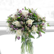 A clear glass cylinder vase holds a poised and peaceful bouquet, illuminated by soft, natural light that could easily be streaming through a Queensbury sitting-room window. At its heart, several large white roses open in flawless spirals, the creamy petals layered so smoothly that they almost appear to glow from within. Intertwined with these roses are multiple stems of alstroemeria, their long, slender petals shaded in cool purples, mauves, and hints of fresh green, adding a gentle veil of colour that never overwhelms the arrangement's calm tone. Tiny clusters of white baby's breath drift among the larger blooms like a fine, airy lace, subtly rounding out the shape of the bouquet and adding a sense of lightness and breath. Lush green foliage-some leaves broad and glossy, others narrow and more delicate-cradles the flowers, creating a structured yet natural frame that evokes the quiet greenery found in local Queensbury gardens and along the nearby streets. The stems are clearly visible through the vase, standing straight in clear water, an honest glimpse of the bouquet's simple, natural construction. Behind the flowers, the background is bright, almost white, with a softly diffused quality that gives the scene a feeling of stillness and reflection, as if captured during a quiet moment before guests arrive for a memorial gathering. The overall impression is one of restraint, purity, and comfort-a dignified arrangement that feels especially fitting for sympathy messages, funeral tributes, or a thoughtful delivery to a grieving family home in and around Queensbury.