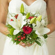 The image shows a romantic bridal bouquet held close to a lace wedding dress, the intricate pattern of the fabric framing the flowers like delicate embroidery against the skin. In the heart of the bouquet, a rich red rose glows with an almost ruby depth, its layered petals suggesting timeless love and devotion. Surrounding it, tall white lilies unfurl in soft, creamy tones, their trumpet-shaped blooms opening with that gentle elegance brides in Queensbury often choose for church weddings or civil ceremonies near Kingsbury and Edgware. Wisps of baby's breath thread through the composition, creating pockets of airy lightness that contrast with the saturated red of the berries dotted among the greenery. Mossy green tufts and textured leaves in multiple shades-from bright, fresh green to deeper woodland hues-add a sense of garden abundance, as if the bouquet had just been gathered from a hidden corner of Fryent Country Park. Fine strands of ornamental grass arch delicately around the flowers, giving the arrangement a sense of movement and softness, like a quiet breath just before the bride steps into the aisle. The bouquet feels lush yet meticulously balanced, each element contributing to an overall mood of refined celebration and heartfelt warmth. It is a hand-tied design that would look at home in any Queensbury wedding venue, radiating romance, freshness, and the intimate thrill of saying I do.