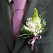 In this detailed image, the camera lingers on a single lapel, telling a whole story about a special day in Queensbury through fabric, colour, and flowers. The wearer's suit is a deep charcoal grey, beautifully tailored, with a smooth, sharply angled lapel that hints at careful preparation and anticipation. Beneath the jacket, a light dress shirt patterned with a fine, dark check peeks through, topped by a lustrous lavender-purple necktie tied in a crisp, traditional knot. The tie's elegant sheen draws the eye upward, where a carefully pinned boutonniere becomes the focal point. White alstroemeria flowers-Peruvian lilies-unfurl with intricate inner markings of reddish-brown and soft yellow, their trumpet-shaped blooms giving both structure and softness. Nestled among them is a tall, tapered green bud, likely tuberose, whose fresh, lime-green hue lifts the arrangement and adds a sense of growth and new beginnings, perfect for weddings or milestone celebrations. Tiny clusters of white gypsophila scatter like stars through the design, offering a cloudlike lightness against the darker suit fabric. The foliage around them mixes slender, spiky stems with feathery greens and broader, polished leaves, creating layers of texture that a florist from Flower Delivery Queensbury might pull straight from an early-morning market run near the local shops. A narrow satin ribbon, dyed a gentle lilac to mirror the tie, is tied in a neat bow at the base of the flowers, firmly anchoring the boutonniere to the lapel while softening it visually. Subtle, even lighting highlights the interplay between cool whites, fresh greens, and rich purples, evoking the calm, hopeful atmosphere of a ceremony taking place not far from Queensbury's tree-lined residential streets.