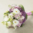 A carefully hand-tied bouquet lies horizontally on a pale beige, lightly textured fabric that could be linen or soft burlap, its understated background highlighting the intricate floral design. The composition is a graceful blend of white, blush pink, and rich purple blooms, all supported by fresh green foliage that threads naturally through the arrangement. White lisianthus flowers take center stage, some open in full, layered rosettes that reveal subtle yellowish-green hearts, while others remain as smooth, rounded buds, adding a sense of unfolding romance. Nestled among them are clusters of stock in shades of soft pink and pale mauve, their many small, ruffled florets forming plush columns of petals that give the bouquet a dreamy, cloudlike quality. To balance the softness, the florist has tucked in striking purple filler flowers, similar to ornamental allium, each tiny star-shaped bloom forming spiky spheres that provide dynamic texture and a bold pop of lavender colour. Long strands of deep green, grass-like foliage curve and loop through the flowers, particularly to the right, adding structure and an elegant, sweeping rhythm that feels as natural as a stroll through Queensbury Park at dusk. The stems are drawn together tightly and finished with a vivid fuchsia satin ribbon, its luxurious sheen enhanced by a fine strand of white pearls that runs along the binding, suggesting quiet luxury and careful craftsmanship. This bouquet, ideal for a bride walking down the aisle in Queensbury or for a refined celebration at home, radiates tenderness, sophistication, and the thoughtful touch of a local florist who understands how to balance texture, colour, and emotion in every stem.