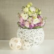 Bathed in the diffused morning light characteristic of Queensbury's welcoming homes, a stunning contemporary bouquet sits atop a luminous white table. The arrangement features plush blossoms of creamy white and pale yellow eustoma, their delicately ruffled petals reminiscent of freshly opened blooms beside Roe Green Park. Vibrant pink matthiola stocks stretch upward, introducing a subtle vertical rhythm and adding a touch of sophistication. Tucked underneath, clusters of deep violet filler flowers-likely aster or dainty allium-contribute a lively, textured base and a gentle fragrance that lingers in the air. All are enfolded by a neat border of deep green leaves, enhancing the organic harmony of the design. The florals reside within a striking white sphere vase, its open-weave shell crafted from painted rattan or fine twigs, giving the piece a light, modern feel. Nestled to the left, two decorative openwork spheres in light rattan mirror the container's artisanal style and infuse the scene with a gentle, tactile warmth. All is set against a creamy beige wall with subtle vertical texture, casting a serene, neutral glow that makes the colours and natural forms truly stand out. This inviting arrangement could grace a spring wedding table, serve as a gentle gesture of celebration, or offer daily delight for anyone in Queensbury seeking artistry and freshness in their floral décor.